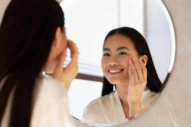 Huzurlu bir sabah ortamında, ipek bornozlu genç Asyalı bir kadın aynanın önünde bir göz bakım ürününü uygularken gülümsüyor. Evde serinletici bir duştan sonra güzellik rutininin tadını çıkarıyor..