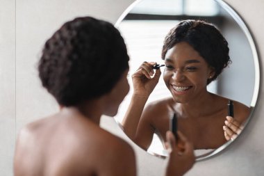 Güzel siyah bir bayan, maskara sürerken, bir aynanın önünde gülümsüyor. Havluyla kaplanarak, hafif aydınlatılmış banyo ortamında makyaj yapmayı sever..