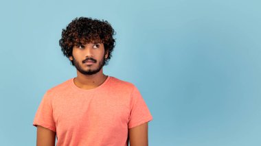 Kıvırcık saçlı genç ve çekici bir Hintli ilginç bir şekilde yana bakıyor. Pembe bir tişört giyer ve mavi arka planlı bir stüdyoda durur. Bir entrika anını yakalar..