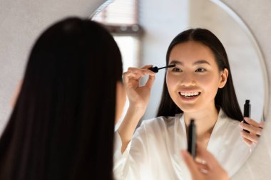 Beyaz bornozlu neşeli genç bir Japon kadın aynanın önünde dururken rimel sürüyor. Makyaj rutinini seviyor, dışarı çıkmaya hazırlanırken gülümsüyor..