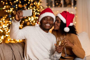 Mutlu genç siyah çift yeni yılı tatil için dekore edilmiş rahat bir odada kutluyor. O selfie çekerken o onu öpüyor. Neşeli kadehlerle dolu kadehlerden içerler..