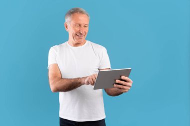 Olgun bir adam dijital tabletle etkileşime girerken gülümser, bir oyuna daldırılır, canlı mavi bir arkaplan üzerine kurulur. Rahatlamış tavrı, teknolojiyle eğlenmeyi ve bağlanmayı öneriyor..