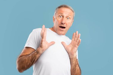 Korkmuş olgun bir adam kameraya geniş gözlerle bakıyor ve mavi bir arka plana karşı savunmacı bir şekilde ellerini kaldırıyor. Yüzündeki ifade korku ve şaşkınlığı gösteriyor, insani duyguların bir anını yakalıyor..