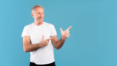 Dostça gülümseyen olgun bir adam heyecan verici bir şekilde kenarı işaret ediyor. Reklamlar için uygun boş bir yer gösteriyor. Mavi bir stüdyoda dikiliyor, dikkat çekmeye hazır..
