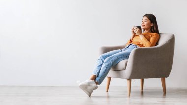 Rahat bir Asyalı kadın rahat bir koltukta oturur, kahvesini yudumlar ve huzurlu bir mola verir. Ayar, dinlenmek için sakin bir atmosfer yaratan parlak beyaz bir duvar içeriyor..