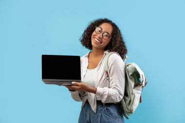 Bin yıllık siyah bir kız öğrenci elinde boş bir ekranla dizüstü bilgisayarını tutarken gülümsüyor. Kıvırcık saçları var, gözlük takıyor ve mavi arka planda duran bir sırt çantasıyla gündelik giyiniyor..