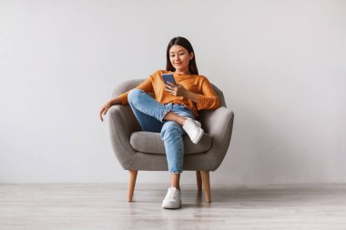 Genç Asyalı kadın rahat bir koltukta rahat rahat akıllı telefonuna bakıyor. Basit bir beyaz stüdyo arkaplanı ile çevrili yeni bir mobil uygulamayı kontrol ederken modern teknolojiyi seviyor..