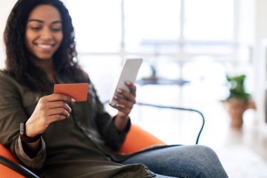 Genç bir kadın modern bir oturma odasında turuncu bir sandalyeye rahatça oturur. Akıllı telefonunu kullanırken ve elinde bir kredi kartı tutarken gülümsüyor. İnternetten alışveriş yaptığını gösteriyor..