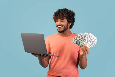 Neşeli genç Hintli bir adam bir elinde modern bir dizüstü bilgisayar, diğer elinde de yüklü miktarda para tutarken gülümsüyor. Mavi bir stüdyo ile çevrimiçi ticaret yapıyor gibi görünüyor..