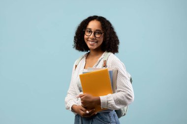 Kendine güvenen siyah bir kadın elinde defterleri ve sırt çantasıyla kameraya gülümsüyor. Akademik eğitim için heyecanını sergileyerek üniversite yolculuğuna başlamaya hazır..