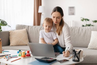 Genç oğlu kucağında otururken bir kadın dizüstü bilgisayarına odaklanır. Renkli oyuncaklar ve okul malzemeleri parlak oturma odalarındaki masanın üzerine yayılmış..