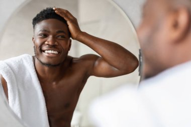 Neşeli bir Afrikalı Amerikalı adam parlak bir banyoda üstsüz duruyor, aynadaki yansımasına bakarken kısa saçlarına dokunuyor. Yeni görünüşüyle mutluluğu ifade ediyor..