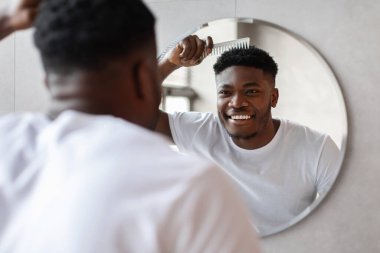 Afrikalı bir Amerikalı aynadaki yansımasına gülümserken kısa saçlarını tarıyor. Şık bir tuvalette, sabah saç stili rutininin tadını çıkarıyor..