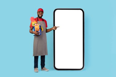 Ev Temizliği Hizmetleri Uygulaması. Siyah, yakışıklı, önlük takan, beyaz ekranlı, büyük, boş, akıllı telefonu işaret eden, elinde sepet dolusu deterjan tutan, gülümseyen, mavi arka plan,