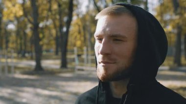 A young man with a beard wearing a hoodie is jogging outdoors. Surrounded by trees, he embraces a healthy lifestyle in a vibrant park. His focus reflects dedication to fitness and well-being.
