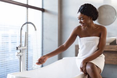 Havluya sarılmış genç siyah bir kadın parlak ve modern bir banyoda akan suyla oynarken gülümsüyor. Rahatlamış görünüyor, rahatlatıcı bir banyo deneyimi için hazır..