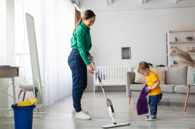 Bir anne, elinde küçük mor bir bez tutarak küçük çocuğu yakınlarda oynarken oturma odasını düzenlemek için bir temizlik aleti kullanır. Güneş ışığı uzayı aydınlatır.