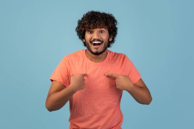 Yakışıklı, bin yıllık Hintli bir adam kendini göstererek şaşkınlığını dile getiriyor. Pembe bir tişört giyiyor ve kıvırcık saçları var. Saf mavi bir arka planı var. Neşeli bir hava katıyor..