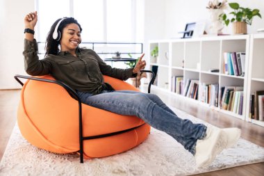 Genç bir kadın rahat ve parlak bir oturma odasında kulaklık takarken hafifçe gülümser ve dans eder. O, kitap rafları ve bitkilerle çevrili bir turuncu fasülye koltuğunda oturuyor..