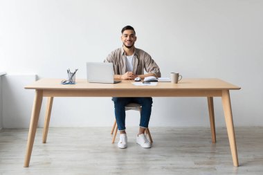 Gülümseyen bir Arap ofis masasında poz veriyor, sıcak bir içeceğin tadını çıkarırken dizüstü bilgisayar kullanıyor. Parlak alan, onun çevredeki iş ve rahatlığına odaklandığını vurguluyor..