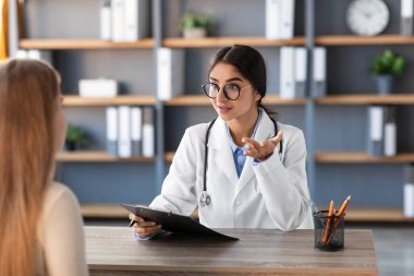 Beyaz önlüklü ve gözlüklü dost canlısı bir Hintli kadın doktor, iyi tasarlanmış bir klinik ofisinde bir müşteriyle istişarede bulunuyor. Atmosfer profesyonel ve misafirperver..