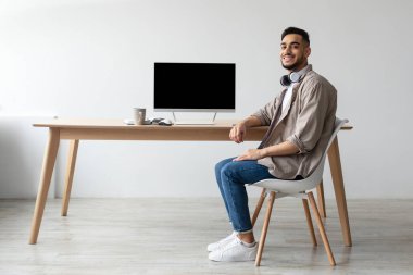 Neşeli bir Arap, ofisinin masasında oturmuş kulaklık takıyor ve gülümsüyor. Boş ekranlı bir dizüstü bilgisayar sergiliyor. Model şablonu için mükemmel..