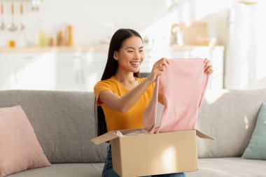A young woman sits on a cozy sofa, smiling broadly as she opens a carton box. She is excitedly checking her delivery of new clothes, enjoying a shopping experience from home.