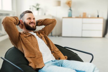 A man with a beard is seated comfortably in a stylish chair, smiling while he leans back with his hands behind his head. The living room is bright and modern.
