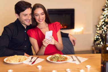 Gülümseyen bir çift makarna tabaklarıyla dolu bir yemek masasında sıcak bir anı paylaşır. Hafif ışıklandırmalı neşeli bir atmosferde telefonlarını birlikte kontrol ediyorlar..