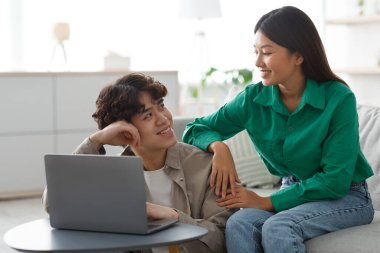 Genç bir Asyalı çift evde dizüstü bilgisayar kullanırken neşeli bir anı paylaşıyor. Çevrimiçi aktivitelere katılırlar, yaşam alanlarında birbirlerine mutluluk ve şefkat gösterirler..