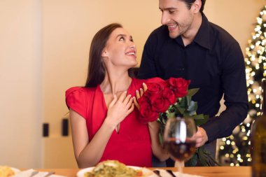 Bir çift romantik bir akşam yemeğinin keyfini çıkarırken bir partner diğerine bir buket kırmızı gülle sürpriz yapıyor. Atmosfer ılık ve şenlikli, yumuşak ışıklandırma ve tatil süslemeleriyle güçlendirilmiş..