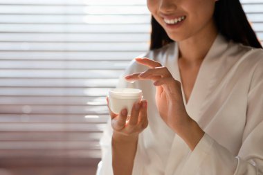 Esmer bir kadının elinde bir kavanoz vücut losyonu tutarken, sabahları nazikçe cildine sürerken. Yumuşak ışık cilt bakımı için rahatlatıcı bir atmosfer yaratır..