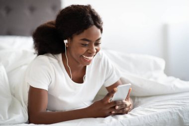 Rahat pijamalı neşeli siyah bir kadın akıllı telefonunu kullanırken yatakta dinleniyor. Müzik dinler ya da arkadaşlarıyla sohbet eder, hafta sonunu evde geçirir..