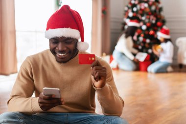 Noel Baba şapkalı neşeli bir siyah adam yerde oturuyor, cep telefonunu Noel hediyesi olarak internetten alışveriş yapıyor. Elinde Noel süsleri ve ailesiyle çevrili bir kredi kartı var..