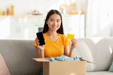 Neşeli bir Koreli kadın elinde boş ekranı ve kredi kartı olan bir akıllı telefonu olan bir paketi kanepeye boşaltıyor. Parlak bir oturma odasında online alışveriş yapmayı seviyor..