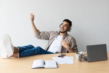 Gülümseyen bir Arap, sanal gitar çalarken, molasının tadını çıkararak evine geri dönüyor. Kablosuz kulaklık takıyor ve ayaklarını masaya dayayıp gevşemeyi ve eğlenceyi somutlaştırıyor..