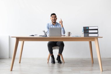 A joyful young business man sits at his desk in a bright office, pointing excitedly upward.