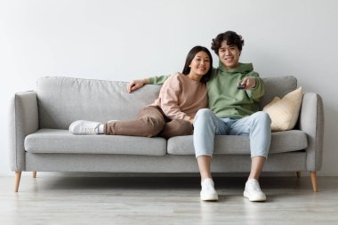 A young Asian couple sits on a couch, cuddling as they watch a movie and use a remote control. They are spending quality time together at home during the weekend.