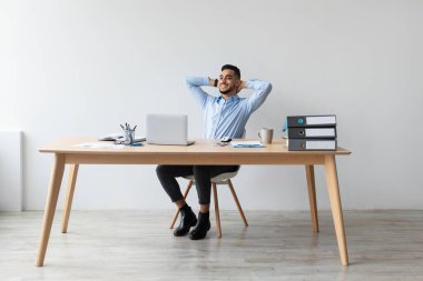 Genç bir Arap adam ahşap masadaki sandalyesine yaslanıyor, gülümsüyor ve rahatlıyor. İşyerinde keyfine bakarken dizüstü bilgisayar kullanıyor, memnun ve memnun görünüyor..