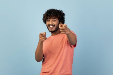 Neşeli genç Hindu adam gülümserken iki eliyle kamerayı işaret ediyor. Mavi bir stüdyo ortamında öylesine poz veriyor, eğlenceli ve çekici bir atmosfer yaratıyor..