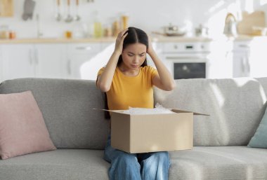 Hayal kırıklığına uğramış bir kadın kanepesinde oturup içinde yanlış eşya olan karton bir kutuya bakıyor. Çevrimiçi alımı ve teslimat hizmeti deneyimi yüzünden üzgün görünüyor..