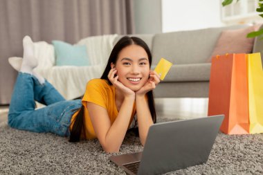 Genç Asyalı bir kadın yumuşak bir halıya uzanmış, dizüstü bilgisayarını internetten kıyafet sipariş etmek için kullanırken gülümsüyor. Elinde renkli alışveriş torbalarıyla dolu bir kredi kartı var. Alışverişin keyfini çıkarıyor..