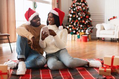 Neşeli bir Afrikalı Amerikalı çift, her ikisi de Noel Baba şapkası takan rahat bir battaniyeye oturur. Hediyelere akıllı telefondan bakıyorlar. Bayram alışverişi için heyecanlılar. Noel süslemeleri etraflarını sarmış..