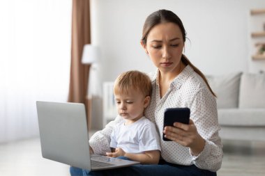 Oturma odasının zemininde oturan bir anne, bebeğinin yanındaki dizüstü bilgisayarla uğraşırken telefonuna odaklanıyor. Oda aydınlık ve davetkar..