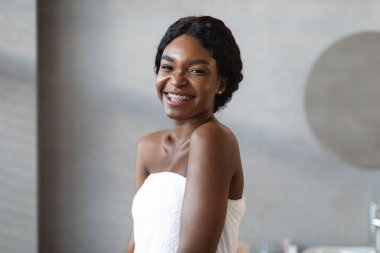Neşeli, Afro-Amerikan bir kadın beyaz bir havluyla modern bir banyoda poz veriyor. Kameraya gülümsüyor, spa gününün ve cilt bakımının keyfini çıkarıyor..