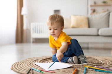 Parlak bir oturma odasında, renkli pastel boyalarla çizime odaklanmış dairesel bir halıya oturan küçük bir çocuk. Yumuşak doğal ışık çevrelerindeki rahat boşluğu vurgular.