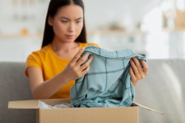 Üzgün Asyalı bir kadın evdeki kanepede oturmuş, bir teslimatı açıyor. Bir paketin üstündeki bluzu incelerken hayal kırıklığına uğramış görünüyor. İnternetteki siparişinden memnun olmadığını gösteriyor..