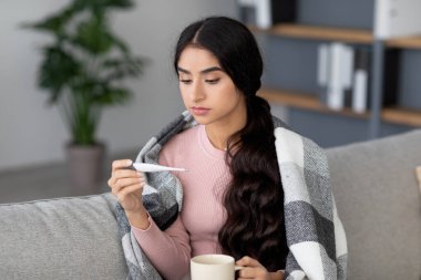 Üzgün genç Hintli kadın kanepede oturur, ekose bir battaniyeye sarınıp termometreye bakar. Bir fincan sıcak içecek tutuyor, grip mevsiminde üşümüş ve hasta hissediyor..