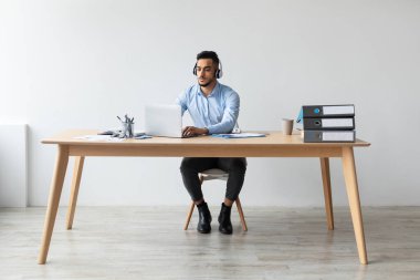 Kulaklık takan bir Arap minimalist bir masada dizüstü bilgisayarına odaklanır. Evrak işleri ve ofis malzemeleriyle modern bir ofiste oturuyor..