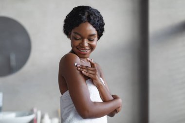 Şehvetli genç kadın omuzlarına vücut losyonu sürerken gülümsüyor. Havluya sarılmış, şık, gri bir banyoda. Serinletici bir duştan sonra kendine bakmanın tadını çıkarıyor..